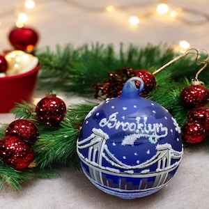 May include: A blue Christmas ornament with the word "Brooklyn" in white script. The ornament features a bridge design and white snowflake accents. Red and gold ornaments and greenery surround the main ornament.