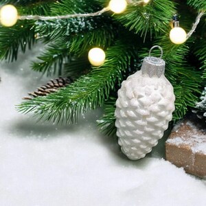 May include: White pine cone shaped Christmas ornament with a silver top hanging from a green pine branch with white fairy lights. The ornament is sitting on a bed of white faux snow.
