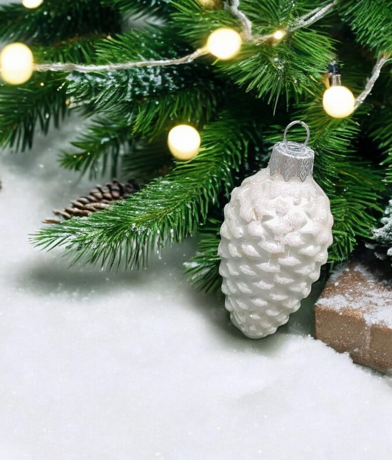 May include: White pine cone shaped Christmas ornament with a silver top hanging from a green pine branch with white fairy lights. The ornament is sitting on a bed of white faux snow.