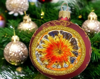 May include: A round, matte burgundy Christmas ornament featuring a central, reflective, starburst-patterned indentation in shades of orange and yellow, surrounded by a textured gold glitter border and delicate gold filigree accents on the surface.