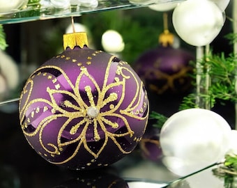 May include: A purple Christmas ornament with a gold floral design. The ornament is sitting on a glass surface with other white ornaments.