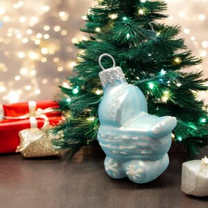 May include: A blue glass Christmas ornament shaped like a baby carriage with white snowflake details. The ornament is hanging from a Christmas tree with white lights.