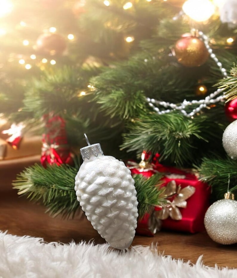 May include: A white pine cone shaped Christmas ornament with a glittery finish hangs on a Christmas tree with red and gold ornaments and white lights.