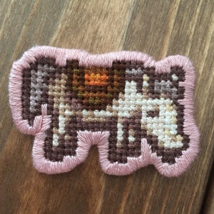 Op de afbeelding: Een roze en bruine geborduurde patch met een gestikt beeld van een koe. De koe is bruin en wit met een bruin en oranje zadel.