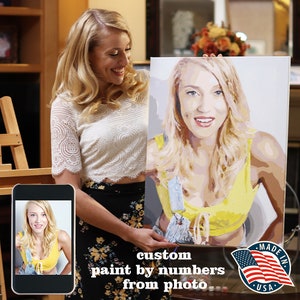 May include: A woman holds up a custom paint by numbers portrait of herself. The portrait is on a canvas and features a yellow top and blue overalls. The text "custom paint by numbers from photo" is below the portrait. The American flag is in the lower right corner with the text "Made in USA".