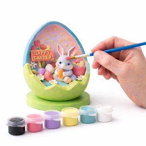 May include: An Easter-themed paint-your-own kit featuring a bunny inside a cracked egg. The egg is light green and blue, with a "Happy Easter" sign. The kit includes paint pots in various colors and a paintbrush.