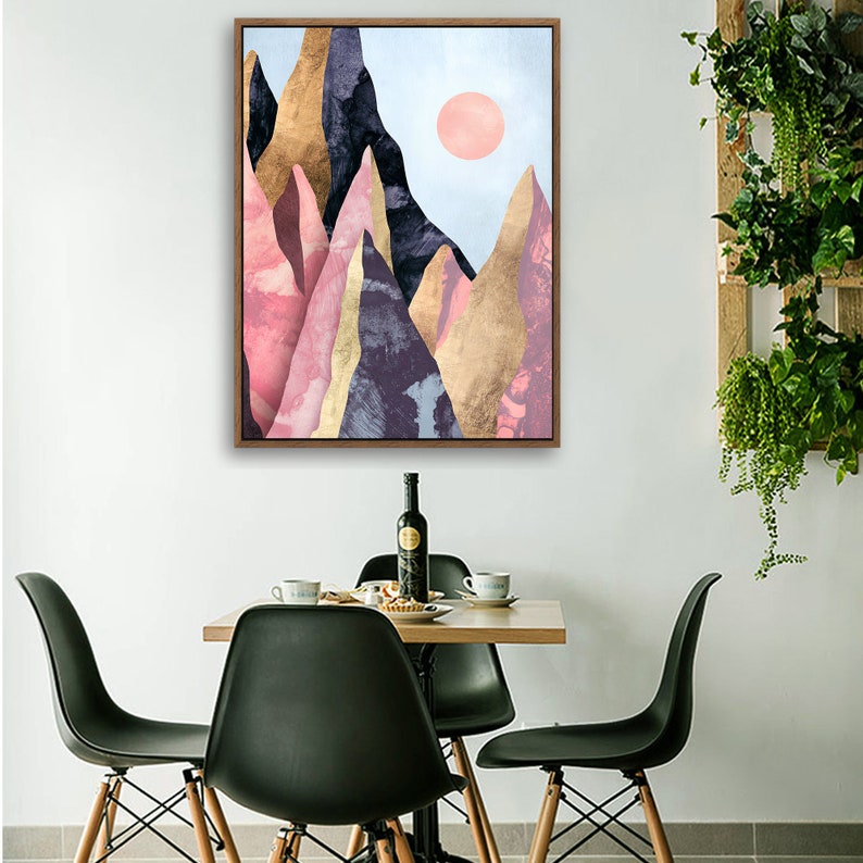 May include: A framed abstract art print featuring a colorful mountain range with a pink sun in the sky. The print is displayed on a wall above a dining table with chairs.