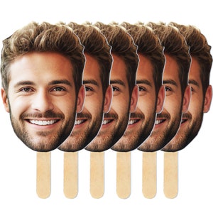 May include: A collection of face cutouts on wooden sticks. Each cutout features a smiling man with light brown hair and a beard. The cutouts are arranged in a row against a white background, suggesting a party or event theme.