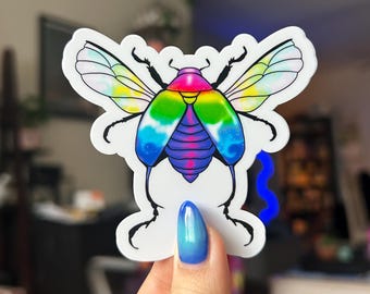 May include: A colorful sticker featuring a stylized insect with rainbow wings and a blue, purple, and pink body. The insect has black legs and antennae. The sticker is held up by a hand with blue nail polish.