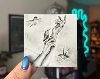 May include: A square, grayscale art print featuring a detailed drawing of hands reaching upwards, with three hummingbirds in flight. The artwork has a soft, textured background, creating a serene and delicate aesthetic.