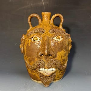 May include: A brown ceramic face jug featuring a sculpted human face with a mustache, goatee, and textured eyebrows. The vessel has two arched handles at the top, a central spout, and visible white teeth set within an open mouth expression.