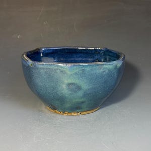 May include: A small, hand-thrown ceramic bowl with a deep blue and teal glazed finish. The bowl features a slightly irregular, faceted rim and a rounded body that tapers down to a visible, unglazed tan clay base at the bottom.