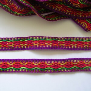 Puede incluir: Borde tejido morado con un patrón geométrico rojo, naranja, verde y dorado. El borde tiene un hilo metálico brillante tejido en el diseño.