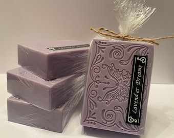 May include: Four bars of lavender scented soap wrapped in plastic and tied with twine. The soap is a light purple color and has a decorative design on the top bar. The label on the top bar reads "Lavender Dreams".