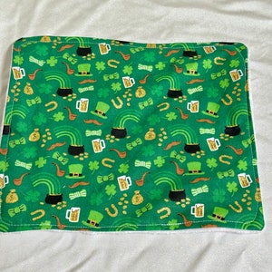 May include: A green fabric square with a white border. The fabric has a repeating pattern of gold coins, shamrocks, rainbows, pipes, leprechaun hats, and bow ties.