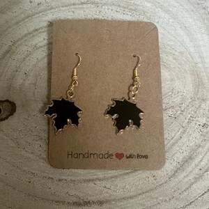 May include: A pair of gold dangle earrings with black leaf-shaped charms. The earrings are on a brown card with the text "Handmade with love".