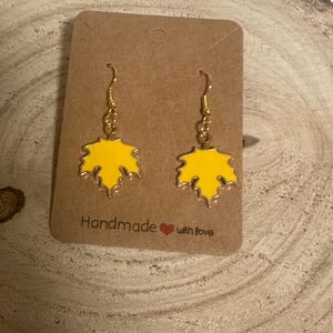 May include: A pair of gold dangle earrings with yellow enamel maple leaf charms. The earrings are displayed on a brown card with the text "Handmade with love".