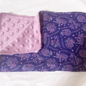 May include: A purple and blue baby blanket with a soft, textured side and a printed side featuring a repeating pattern of mythical creatures and the text "Ravenclaw, Hufflepuff, Slytherin, Gryffindor".
