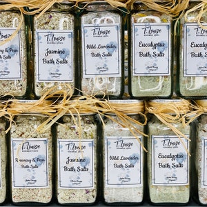 May include: A collection of glass jars filled with bath salts. The jars are labeled with different scents, including Rosemary & Pear, Jasmine, Wild Lavender, and Eucalyptus. The jars are arranged in two rows, with the top row slightly offset from the bottom row.
