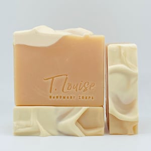 May include: Two bars of handmade soap, one is a light peach color with a white swirl on top and the other is a white bar with a peach swirl. The peach bar has the text "T. Louise Handmade Soaps" printed on the front.