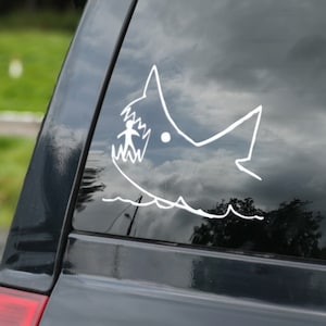 May include: A black car with a white shark sticker on the rear window. The shark is drawn in a simple style and has a person inside its mouth.