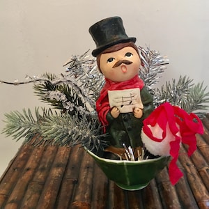 May include: A vintage ceramic figurine of a singing man wearing a black top hat, red scarf, and green coat. He holds sheet music and is surrounded by silver tinsel and evergreen branches. A red and white Santa hat is nearby.