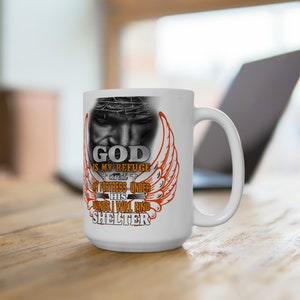 May include: White ceramic coffee mug with a design of Jesus Christ with a crown of thorns and wings. The text reads "God is my refuge and my fortress under his wings I will find shelter".