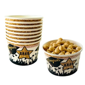 May include: A stack of 10 gold glitter graduation party cups with black silhouettes of people wearing graduation caps and the words "Congrats Grad". One cup is filled with roasted hazelnuts.