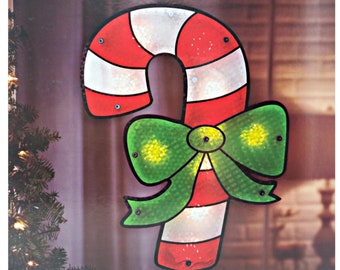 Puede incluir: Un bastón de caramelo rojo y blanco con un lazo verde, iluminado con luces. El bastón de caramelo es una decoración navideña.