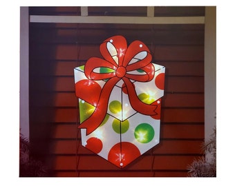 Puede incluir: Una caja de regalo blanca con lunares rojos y verdes y un lazo rojo. La caja está iluminada con luces blancas.