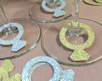 May include: Two wine glasses with gold and silver glitter ring designs on the bottom. There are also two gold glitter ring designs on a white surface.