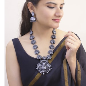 May include: A silver-toned necklace and matching earrings set. The necklace features a pendant with a central figure and smaller figures along the chain. The earrings are bell-shaped. The jewelry is worn with a black top.