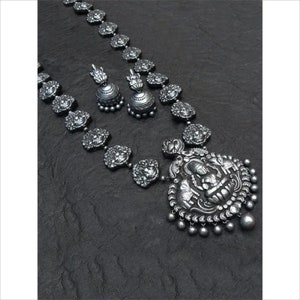 May include: A silver-toned necklace and earring set. The necklace features a large pendant with a detailed figure and small dangling beads. The chain is adorned with ornate, circular elements. The matching earrings have a bell shape with a decorative top.