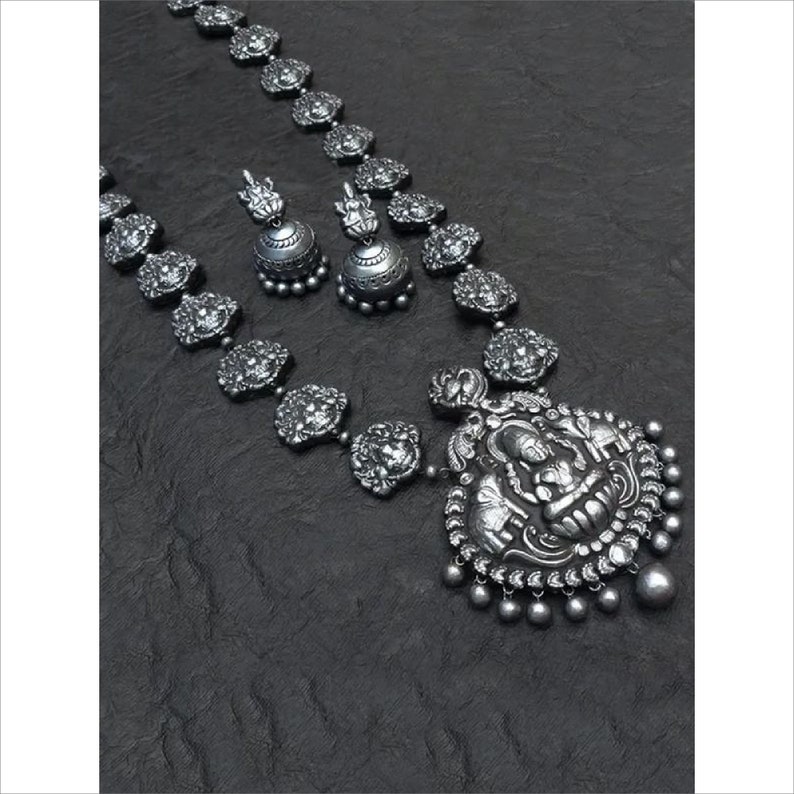 May include: A silver-toned necklace and earring set. The necklace features a large pendant with a detailed figure and small dangling beads. The chain is adorned with ornate, circular elements. The matching earrings have a bell shape with a decorative top.