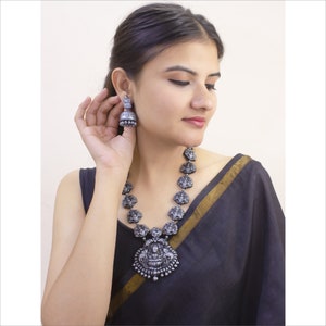 May include: A silver-toned necklace and matching earrings set. The necklace features a large pendant with intricate carvings and smaller, detailed pendants. The earrings are bell-shaped. The jewelry is worn with a black garment.