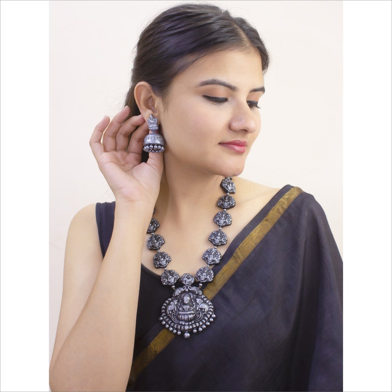 May include: A silver-toned necklace and matching earrings set. The necklace features a large pendant with intricate carvings and smaller, detailed pendants. The earrings are bell-shaped. The jewelry is worn with a black garment.