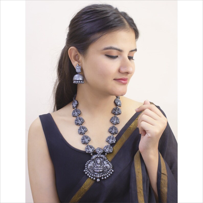 May include: A silver-toned necklace and matching earrings set. The necklace features a pendant with a central figure and smaller figures along the chain. The earrings are bell-shaped. The jewelry is worn with a black top.