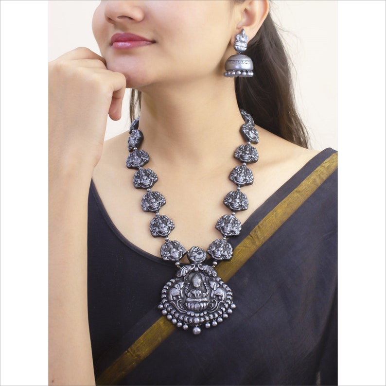 May include: A silver-toned necklace and matching earrings set. The necklace features a pendant with a seated figure and a series of smaller, detailed figures. The earrings are bell-shaped. The jewelry is worn over a black garment.