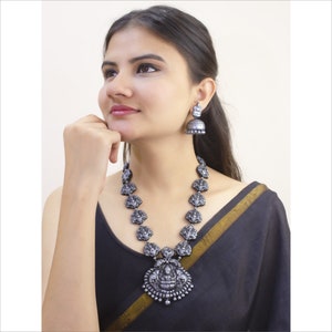 May include: A silver-toned necklace and matching earrings set. The necklace features a long chain with detailed pendant designs and a larger central pendant. The earrings have a bell-shaped design. The jewelry is worn against a black garment.