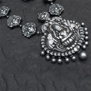 May include: Silver-toned necklace featuring a detailed pendant with an embossed design of a seated figure and elephants. The necklace has a series of round, decorative elements and small dangling beads. The background is a dark, textured surface.