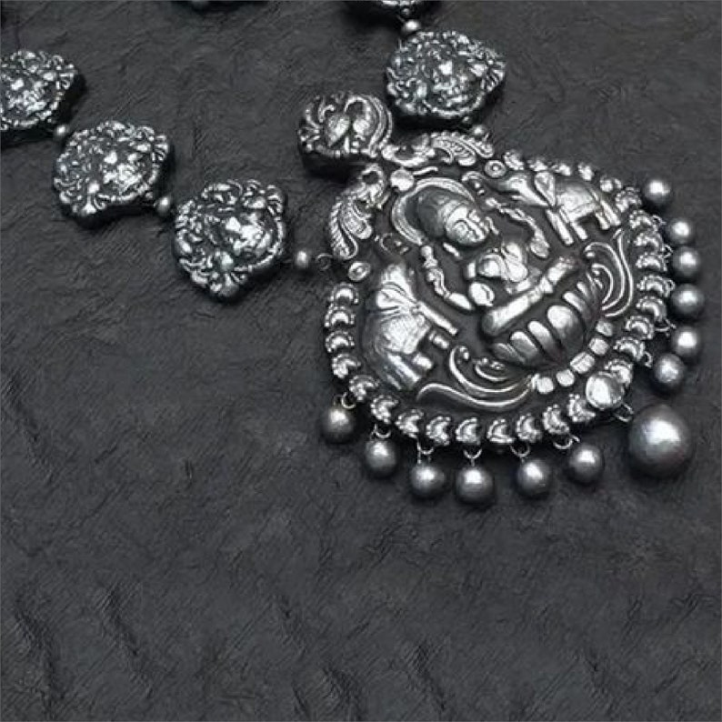 May include: Silver-toned necklace featuring a detailed pendant with an embossed design of a seated figure and elephants. The necklace has a series of round, decorative elements and small dangling beads. The background is a dark, textured surface.