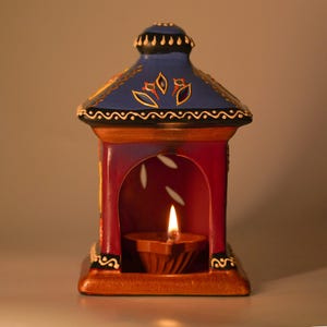 Puede incluir: Una linterna decorativa de cerámica pintada a mano en tonos rojos, azules y dorados. La linterna presenta una pequeña lámpara de aceite con una llama encendida en su interior. El diseño incluye patrones florales y un techo estilo pagoda.