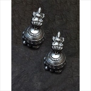 May include: A pair of silver-toned earrings. Each earring features a detailed figure atop a bell-shaped base. The base is adorned with intricate patterns and small, round embellishments. The earrings are displayed against a dark, textured background.