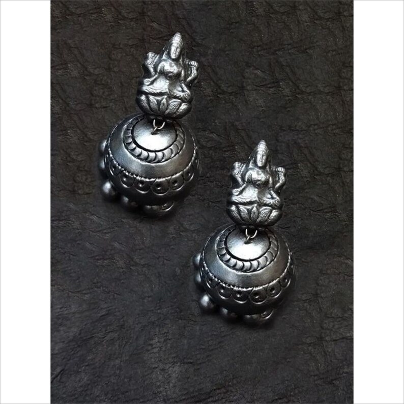 May include: A pair of silver-toned earrings. Each earring features a detailed figure atop a bell-shaped base. The base is adorned with intricate patterns and small, round embellishments. The earrings are displayed against a dark, textured background.