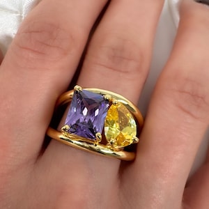 May include: A gold ring featuring a large, square, purple gemstone and a smaller, pear-shaped, yellow gemstone. The ring has a thick, double-band design. The gemstones are set in a gold band.