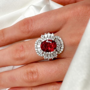 Könnte beinhalten: Ein Ring mit einem großen ovalen roten Edelstein, der von kleineren weißen Edelsteinen umgeben ist. Der Ring ist in einer silbernen Fassung gefasst.