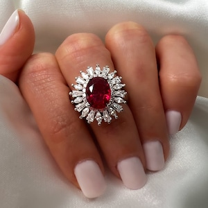 May include: A silver ring with a large red oval gemstone surrounded by smaller clear gemstones.