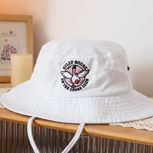 Può includere: Cappello da pescatore bianco con un logo ricamato rosa e nero che dice "Silly Goose on the Loose Club".