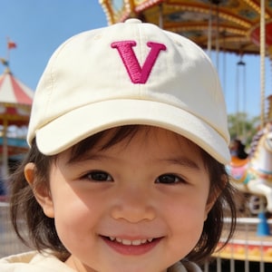 May include: A young child wearing a cream-colored baseball cap with a magenta letter "V" embroidered on the front. The child is also wearing a cream-colored zip-up hoodie. A carousel is visible in the background.