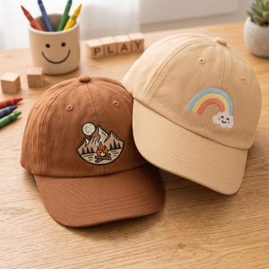 以下が含まれることがあります： 山とキャンプファイヤーの刺繍が施されたミディアムブラウンの野球帽と、虹と笑顔の雲の刺繍が施されたライトベージュの野球帽の2つ。どちらの帽子も、しっかりとしたクラウンとカーブしたつばが特徴です。